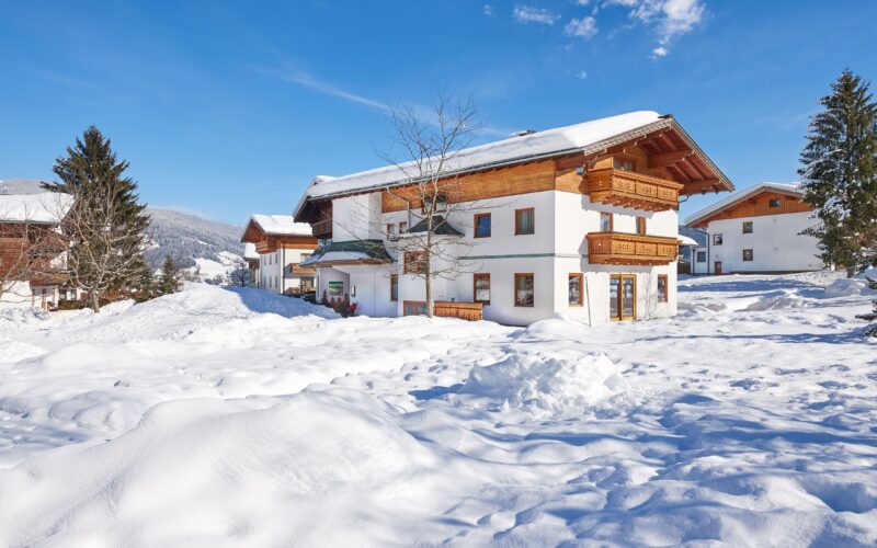 Ein Haus im Chalet-Stil mit Holzbalkonen liegt inmitten von tiefem Schnee unter einem klaren blauen Himmel, mit Bergen und Bäumen im Hintergrund.