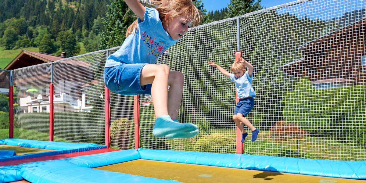 Zwei Kinder springen auf Trampolinen im Freien, die von Sicherheitsnetzen umgeben sind. Im Hintergrund sind an einem sonnigen Tag Bäume und Berge zu sehen.