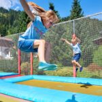 Zwei Kinder springen auf Trampolinen im Freien, die von Sicherheitsnetzen umgeben sind. Im Hintergrund sind an einem sonnigen Tag Bäume und Berge zu sehen.