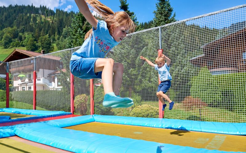 Zwei Kinder springen auf Trampolinen im Freien, die von Sicherheitsnetzen umgeben sind. Im Hintergrund sind an einem sonnigen Tag Bäume und Berge zu sehen.