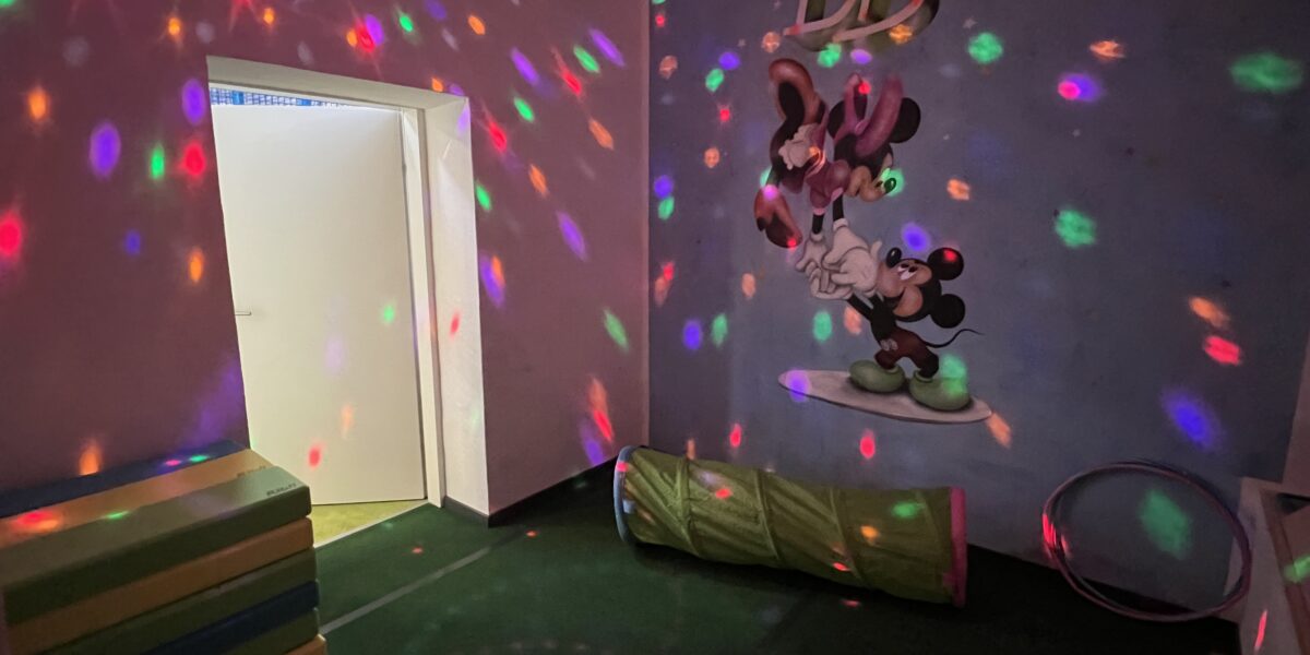 Ein kleiner Raum mit bunten Discolichtern, Schaumstoffmatten, einem Spieltunnel, einem Hula-Hoop-Reifen und einem Wandgemälde mit Cartoon-Mäusen unter dem Wort "DISCO".