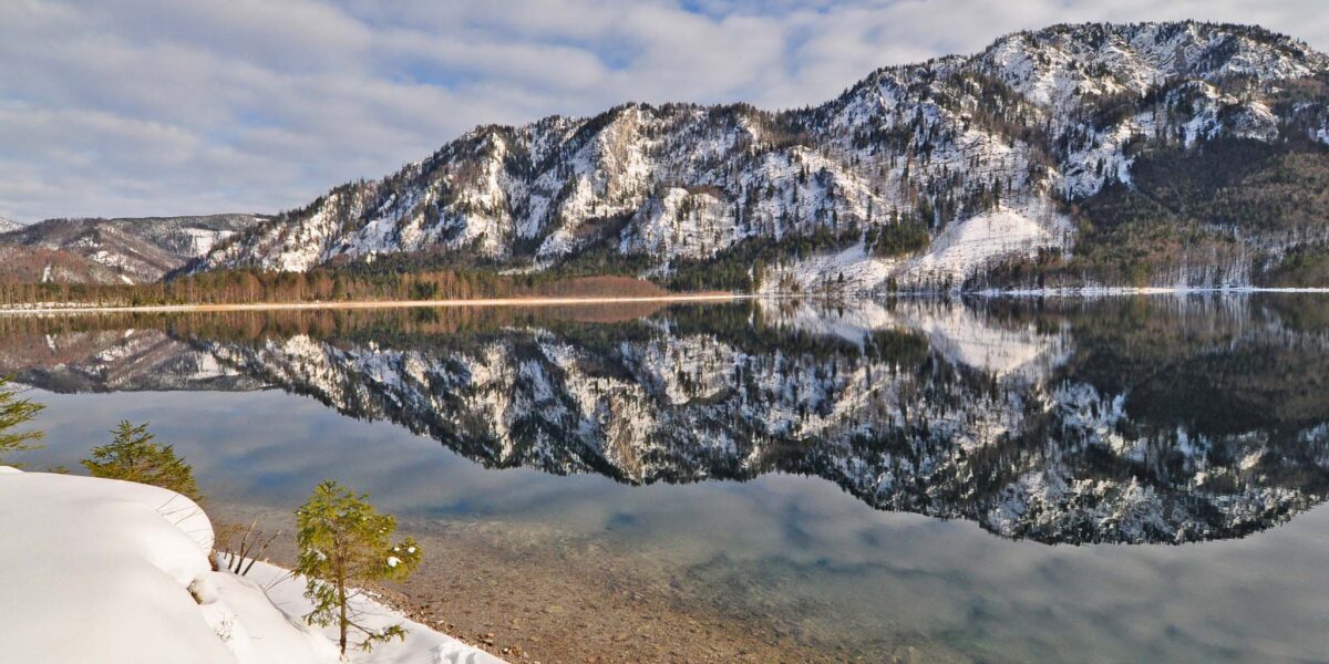 Schneebedeckte Berge und Bäume spiegeln sich in einem ruhigen, klaren See unter einem teilweise bewölkten Himmel, wobei der Schnee die Uferlinie im Vordergrund bedeckt.