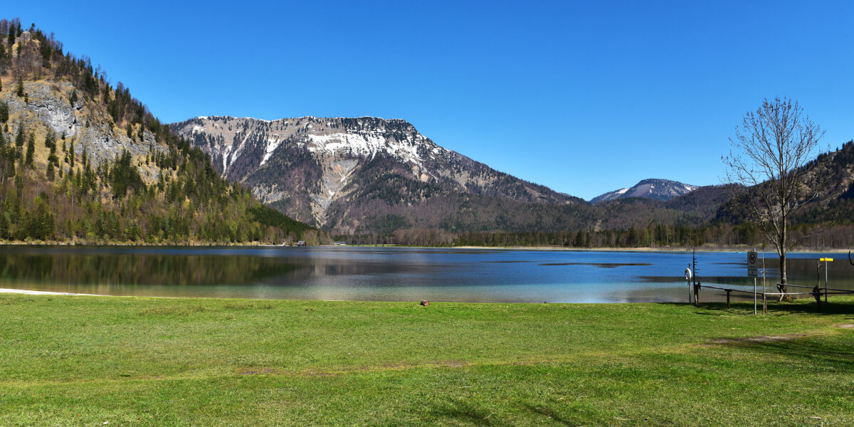 Ein klarer See mit Spiegelungen, grasbewachsenem Ufer und schneebedeckten Bergen unter blauem Himmel; rechts stehen zwei Personen auf einem Holzsteg.