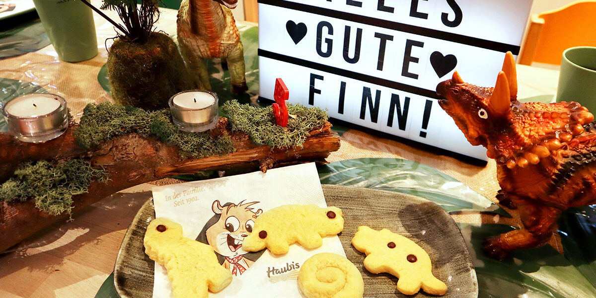 Ein Teller mit Keksen in Form von Dinosauriern auf einem Tisch mit Spielzeugdinosauriern, Kerzen und einem Schild mit der Aufschrift "ALLES GUTE FINN" im Hintergrund.