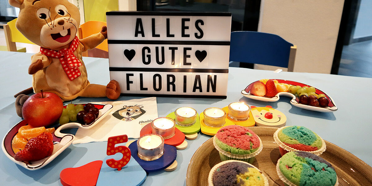 Ein Kindergeburtstagstisch mit einem Teddybär, einer Schrifttafel mit der Aufschrift "ALLES GUTE FLORIAN", Obst, bunten Muffins, Kerzen und einer hölzernen Zahl 5.