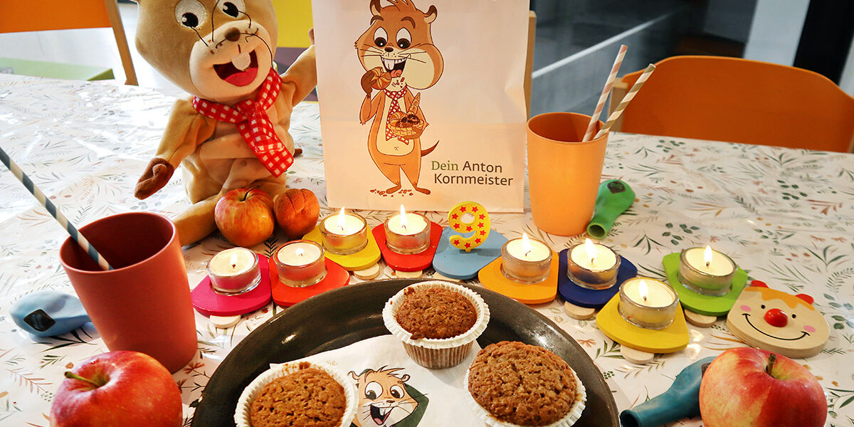Ein Tisch mit Muffins, Äpfeln, Tassen, brennenden Kerzen, einem Plüschhamster und einer Geschenktüte mit einem Cartoon-Hamster und deutschem Text, bereit für eine Feier.