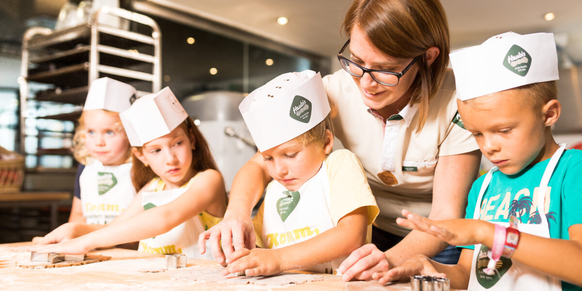 Ein Erwachsener hilft Kindern, an einem bemehlten Tisch in einer hell erleuchteten Küche mit Ausstechern Teig zu formen. Alle tragen Kochmützen und Schürzen aus Papier.