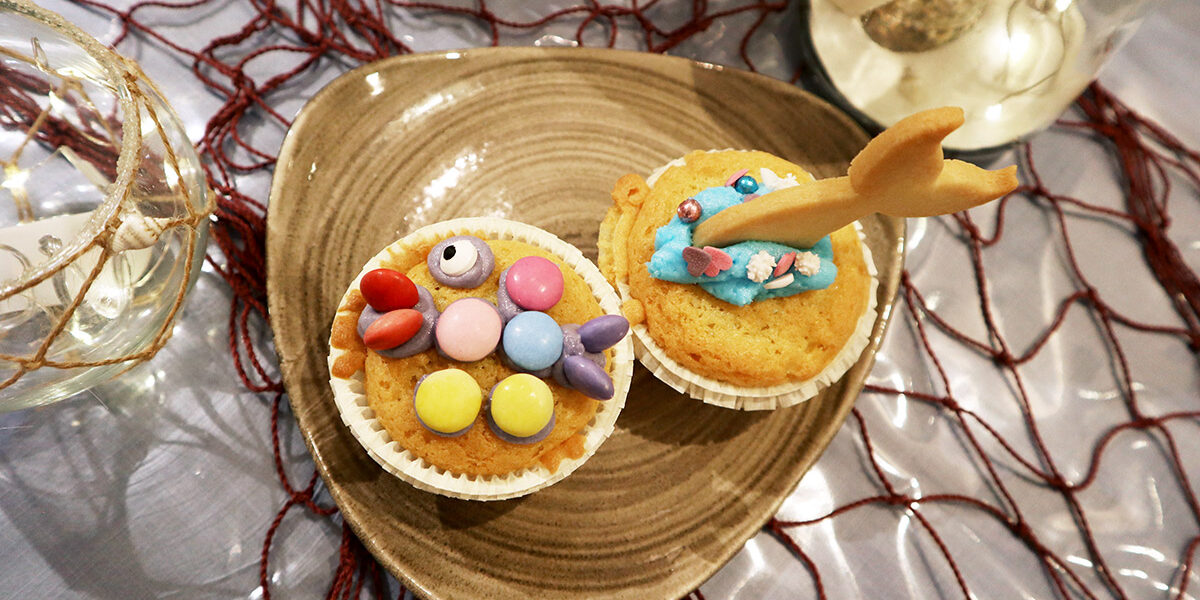 Zwei dekorierte Cupcakes auf einem hellbraunen Teller; der eine hat bunte Süßigkeiten in Form eines Fisches, der andere blaue Glasur und einen Keks in Form eines Meerjungfrauenschwanzes.
