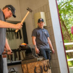 Ein Schmied schlägt erhitztes Metall auf einen Amboss, während zwei Männer, einer in Arbeitskleidung und einer in einem rosa Hemd, in einer offenen Werkstatt beobachten.