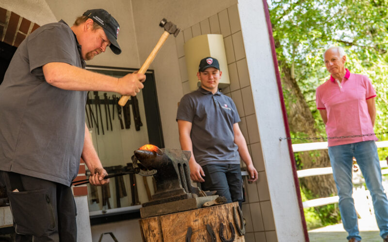 Ein Schmied schlägt erhitztes Metall auf einen Amboss, während zwei Männer, einer in Arbeitskleidung und einer in einem rosa Hemd, in einer offenen Werkstatt beobachten.