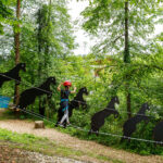 Eine Person mit Sicherheitsausrüstung überquert eine mit schwarzen Pferdesilhouetten geschmückte Seilbrücke in einem bewaldeten Outdoor-Abenteuerpark.