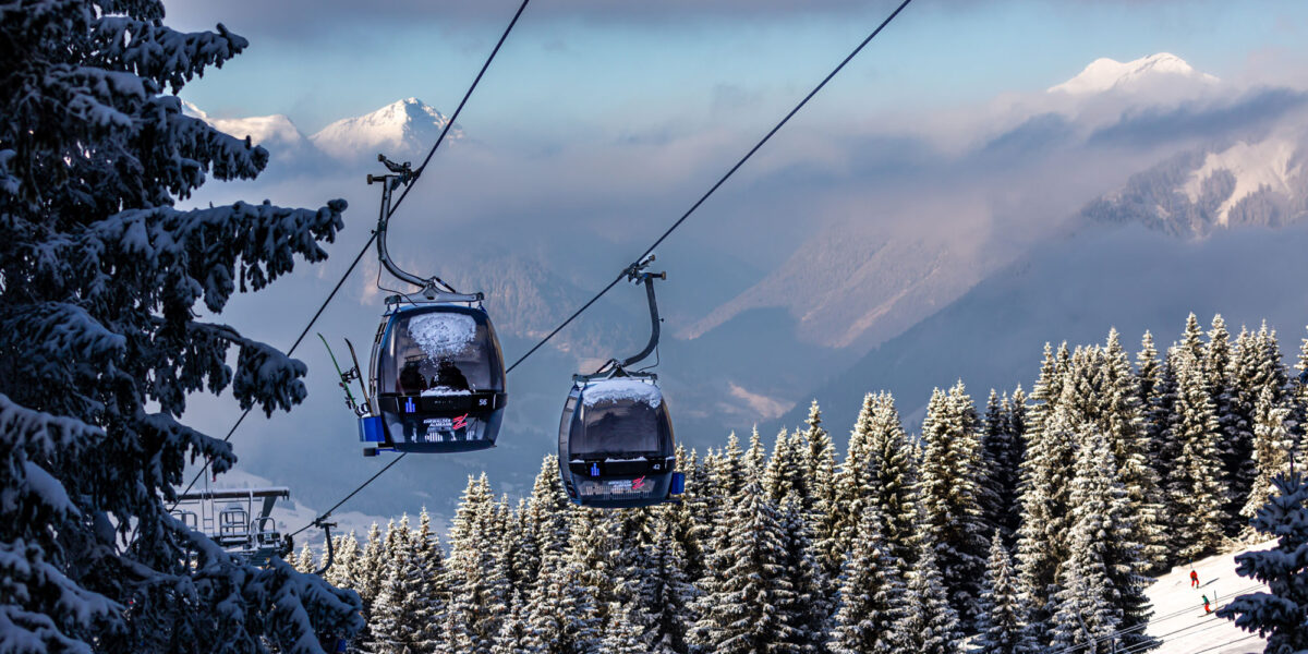 Zwei Skilifte befördern Personen über einen verschneiten, bewaldeten Berghang mit schneebedeckten Bäumen und fernen Bergen im Hintergrund.