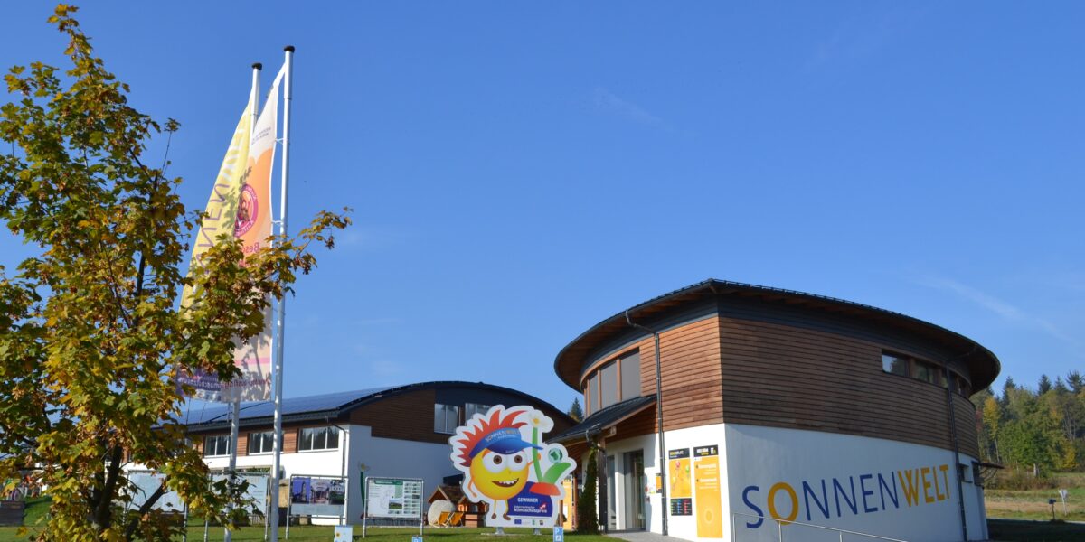 Ein modernes, rundes Gebäude mit dem Schriftzug "SONNENWELT", zwei großen Fahnen, einem Baum und einem fröhlichen Schild mit einer Comicfigur am Eingang unter einem strahlend blauen Himmel.