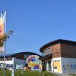 Ein modernes, rundes Gebäude mit dem Schriftzug "SONNENWELT", zwei großen Fahnen, einem Baum und einem fröhlichen Schild mit einer Comicfigur am Eingang unter einem strahlend blauen Himmel.