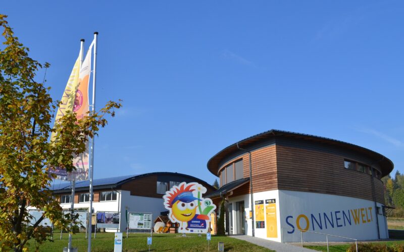 Ein modernes, rundes Gebäude mit dem Schriftzug "SONNENWELT", zwei großen Fahnen, einem Baum und einem fröhlichen Schild mit einer Comicfigur am Eingang unter einem strahlend blauen Himmel.