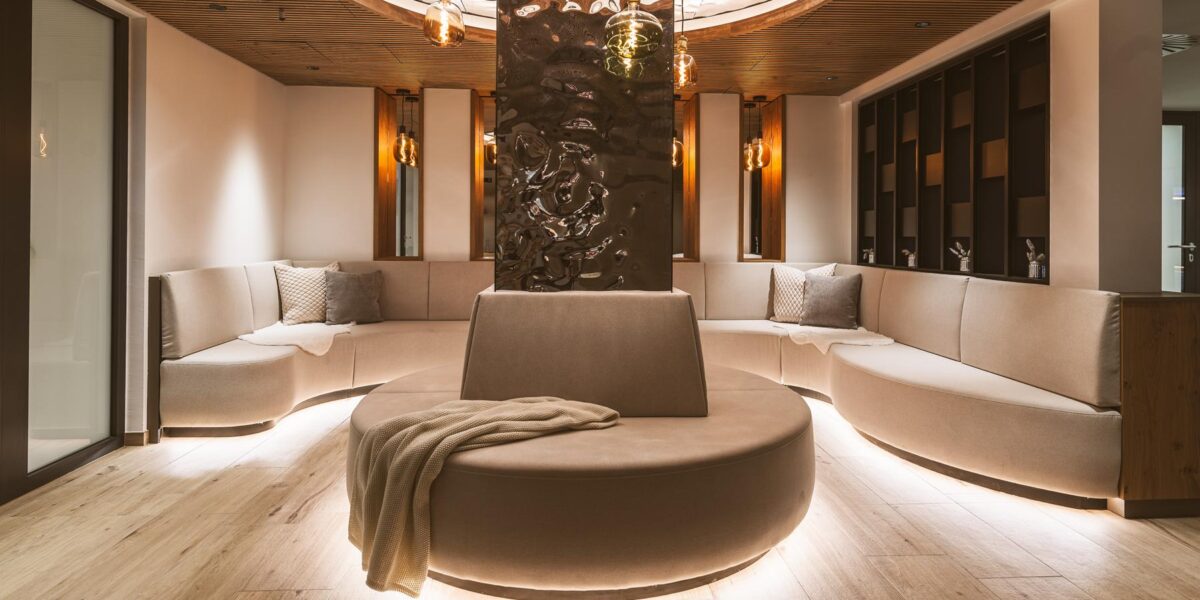 Moderner Lounge-Bereich mit geschwungenen beigen Sofas, einem runden, mit einer Decke drapierten Ottomanen in der Mitte, sanfter Raumbeleuchtung und Holzakzenten an Decke und Boden.