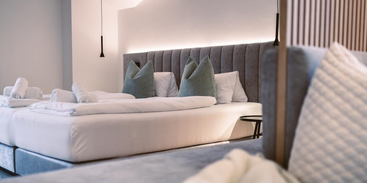 Modernes Hotelzimmer mit sauber gemachtem Doppelbett, grauem Polsterkopfteil, grünen Akzentkissen, aufgerollten weißen Handtüchern und sanfter Raumbeleuchtung.