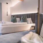 Modernes Hotelzimmer mit sauber gemachtem Doppelbett, grauem Polsterkopfteil, grünen Akzentkissen, aufgerollten weißen Handtüchern und sanfter Raumbeleuchtung.