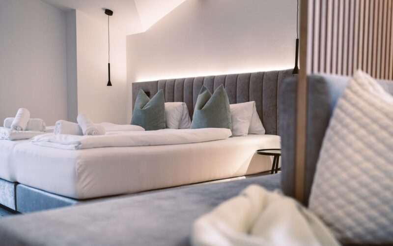 Modernes Hotelzimmer mit sauber gemachtem Doppelbett, grauem Polsterkopfteil, grünen Akzentkissen, aufgerollten weißen Handtüchern und sanfter Raumbeleuchtung.
