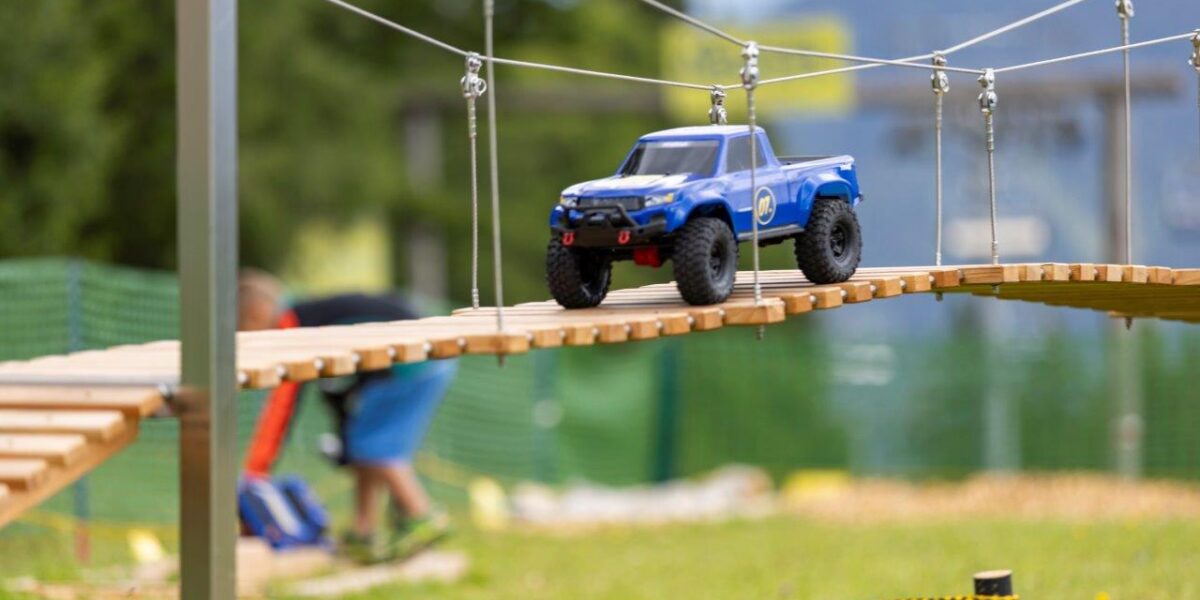 Ein blauer, ferngesteuerter Lastwagen fährt über eine hängende Holzbrücke im Freien, mit unscharfen Menschen und Grünpflanzen im Hintergrund.