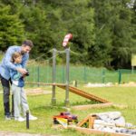 Ein Erwachsener hilft einem Kind, ein ferngesteuertes Auto auf einem von Gras und Bäumen umgebenen Hindernisparcours zu steuern.