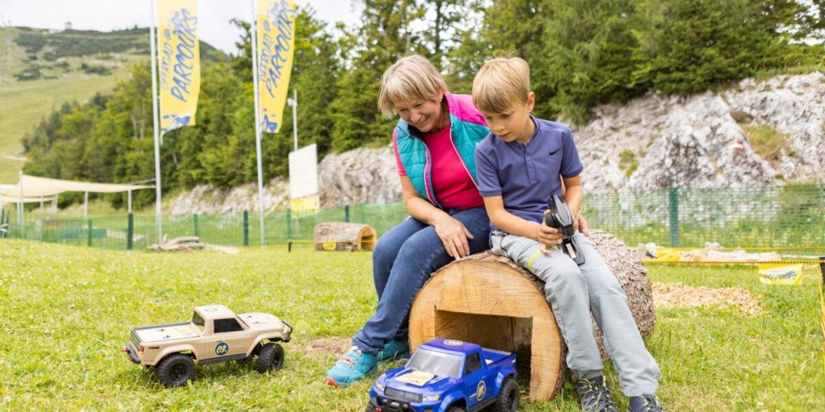 Eine Frau und ein Junge sitzen auf einem Baumstamm im Freien und bedienen zwei ferngesteuerte Spielzeuglastwagen auf dem Gras, mit Bäumen und Bergen im Hintergrund.