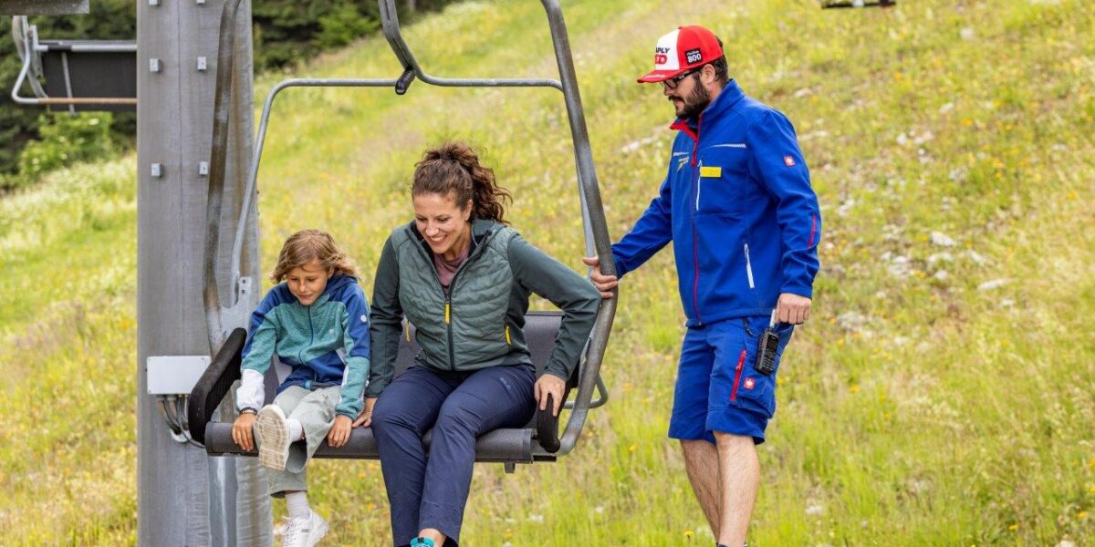 Ein Kind und ein Erwachsener sitzen auf einem Skiliftsessel, während ein Begleiter in blauer Uniform ihnen am Einstiegsbereich mit grasbewachsenen Pisten im Hintergrund hilft.