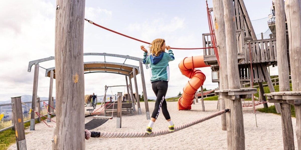 Ein Kind balanciert auf einer Seilbrücke auf einem Spielplatz im Freien, umgeben von Holzstrukturen und einer roten Spiralrutsche.
