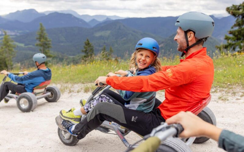 Zwei Personen mit Helmen fahren mit einem Go-Kart auf einem Schotterweg mit Bergen und Bäumen im Hintergrund. Ein weiterer Fahrer ist in der Ferne zu sehen.
