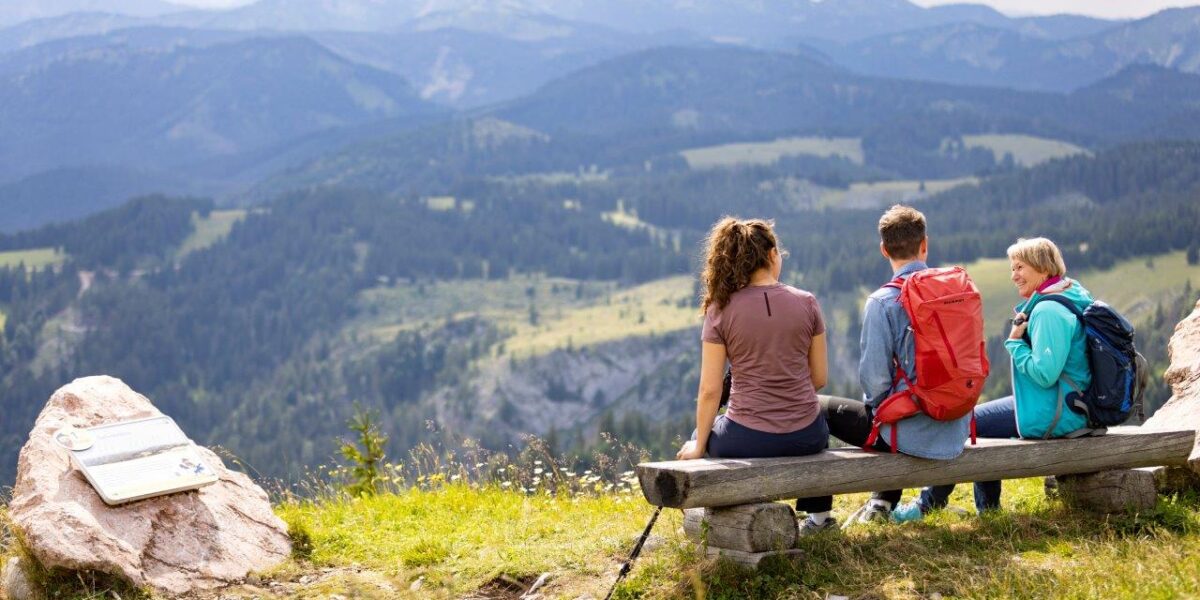 Drei Personen sitzen auf einer Holzbank mit Blick auf eine Berglandschaft, neben ihnen eine Wanderausrüstung und in der Nähe auf einem Felsen ein Wegweiser.