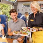 Ein Kellner in einer gelben Schürze reicht einem Mann ein Tablett mit Essen, während in einem modernen Speisewagen ein Kind neben ihm lächelt.