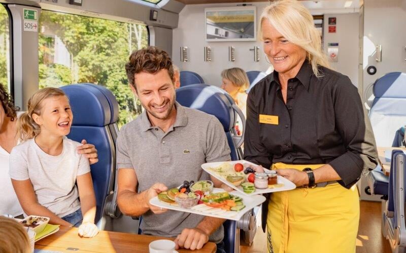 Ein Kellner in einer gelben Schürze reicht einem Mann ein Tablett mit Essen, während in einem modernen Speisewagen ein Kind neben ihm lächelt.