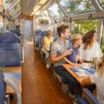 Die Passagiere sitzen an Tischen und genießen Speisen und Getränke in einem modernen Zugwaggon mit großen Fenstern und einem Glasdach, draußen umgeben von grünen Bäumen.