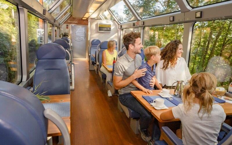 Die Passagiere sitzen an Tischen und genießen Speisen und Getränke in einem modernen Zugwaggon mit großen Fenstern und einem Glasdach, draußen umgeben von grünen Bäumen.