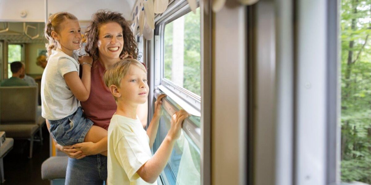 Eine Frau und zwei Kinder schauen aus dem Fenster eines fahrenden Zuges. Die Frau hält ein Kind, während das andere steht. Draußen sind Bäume zu sehen.