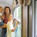 Eine Frau und zwei Kinder schauen aus dem Fenster eines fahrenden Zuges. Die Frau hält ein Kind, während das andere steht. Draußen sind Bäume zu sehen.