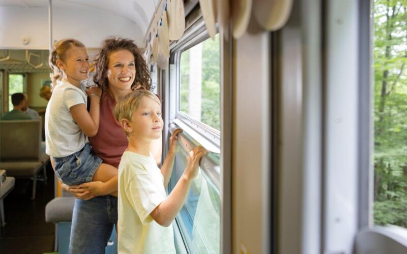 Eine Frau und zwei Kinder schauen aus dem Fenster eines fahrenden Zuges. Die Frau hält ein Kind, während das andere steht. Draußen sind Bäume zu sehen.