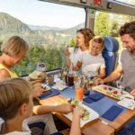 Eine fünfköpfige Familie genießt eine gemeinsame Mahlzeit an einem Tisch in einem modernen Zug mit großen Fenstern und Blick auf eine malerische Berglandschaft.