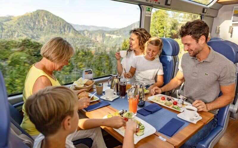 Eine fünfköpfige Familie genießt eine gemeinsame Mahlzeit an einem Tisch in einem modernen Zug mit großen Fenstern und Blick auf eine malerische Berglandschaft.