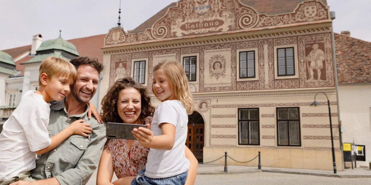 Eine vierköpfige Familie macht ein Selfie vor einem historischen Gebäude mit kunstvollen Verzierungen und dem Wort "Rathaus" an der Fassade.