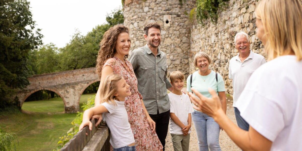 Eine Gruppe von sechs Personen, darunter zwei Kinder, steht lächelnd in der Nähe einer Steinmauer und einer Brücke, während eine Frau im Vordergrund im Freien zu ihnen spricht.