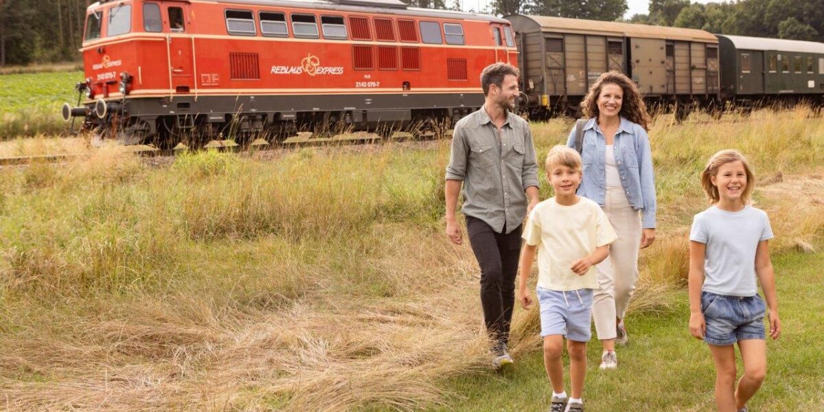 Eine vierköpfige Familie geht auf einer Wiese in der Nähe eines roten Zuges spazieren, mit Bäumen und Waggons im Hintergrund.
