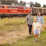 Eine vierköpfige Familie geht auf einer Wiese in der Nähe eines roten Zuges spazieren, mit Bäumen und Waggons im Hintergrund.