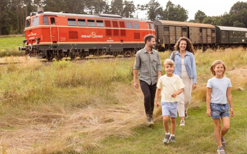 Eine vierköpfige Familie geht auf einer Wiese in der Nähe eines roten Zuges spazieren, mit Bäumen und Waggons im Hintergrund.