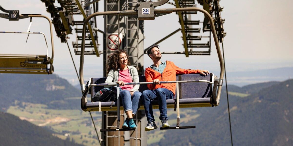 Zwei Personen sitzen in einem offenen Sessellift, der über eine bergige Landschaft hinauffährt. Im Hintergrund sind der klare Himmel und die fernen Berge zu sehen.