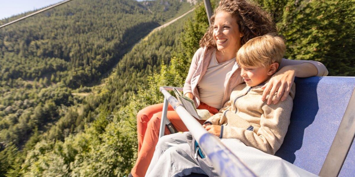 Eine Frau und ein Kind sitzen zusammen in einem blauen Sessellift, der an einem sonnigen Tag über eine bewaldete Berglandschaft hinauffährt.