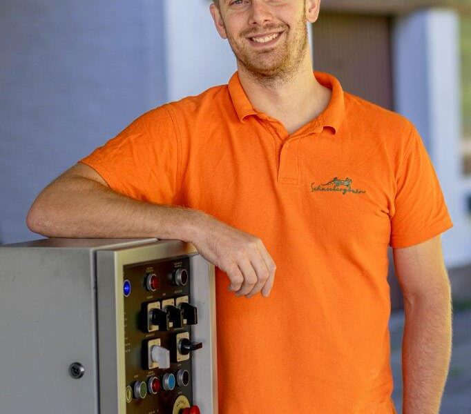 Ein Mann in einem orangefarbenen Poloshirt steht im Freien neben einer industriellen Schalttafel und lächelt in die Kamera.