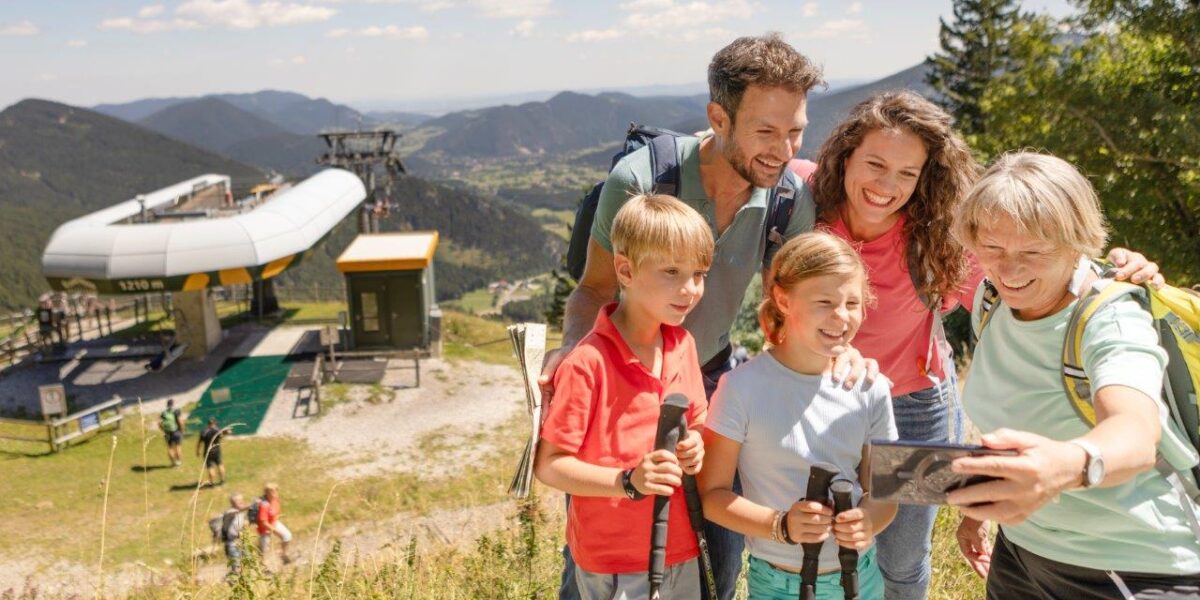 Eine Gruppe von fünf Personen mit Wanderausrüstung macht ein Selfie auf einem Bergpfad in der Nähe eines Skilifts, mit einem malerischen Tal und bewaldeten Hügeln im Hintergrund.