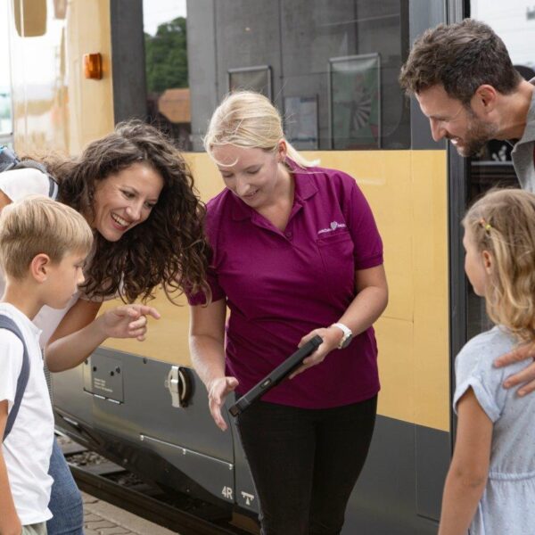 Ein Bahnmitarbeiter zeigt einer lächelnden Familie mit zwei Kindern, die auf einem Bahnsteig neben einem gelben Zug steht, Informationen auf einem Tablet.