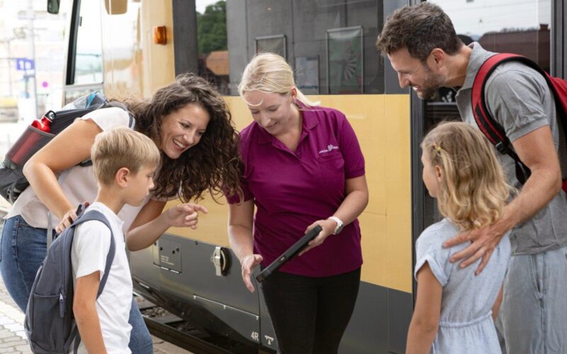 Ein Bahnmitarbeiter zeigt einer lächelnden Familie mit zwei Kindern, die auf einem Bahnsteig neben einem gelben Zug steht, Informationen auf einem Tablet.